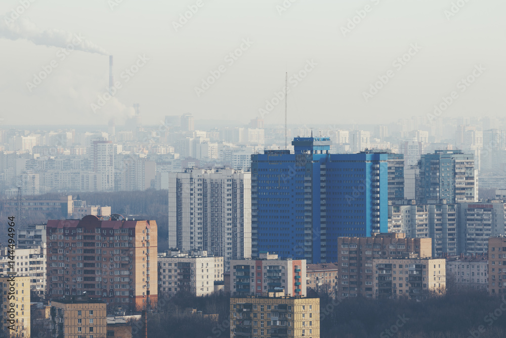 Close-up shooting from high point of residential district in metropolitan city with tall blue building, multiple residential houses of different sizes, bare trees on winter day and smoking chimneys
