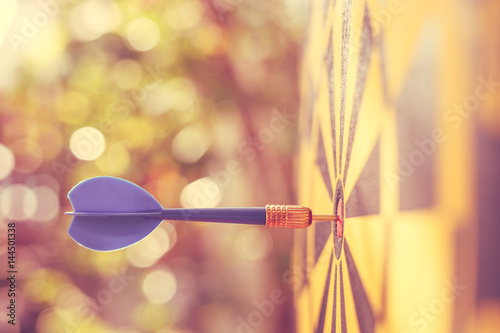 Blue dart arrow in the center of dartboard. Blur and bokeh in sunrise time background. Warm vintage effect style