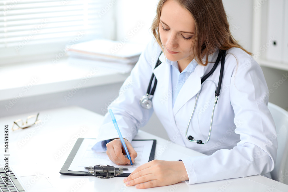 Young female doctor filling up medical form while sitting at the table near window in hospital Young female doctor filling up medical form while sitting at the table near window in hospital