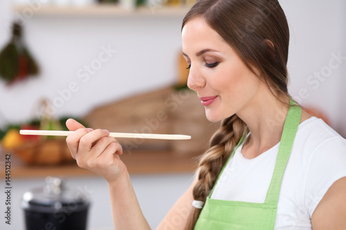 Young woman is cooking in a kitchen. Housewife is tasting the soup by wooden spoon.