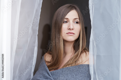 Close up shot of attractive lady with long hair looking outside her bedroom after waking up. Tender young woman is wearing nothing but warm gray blanket on her shoulders.
