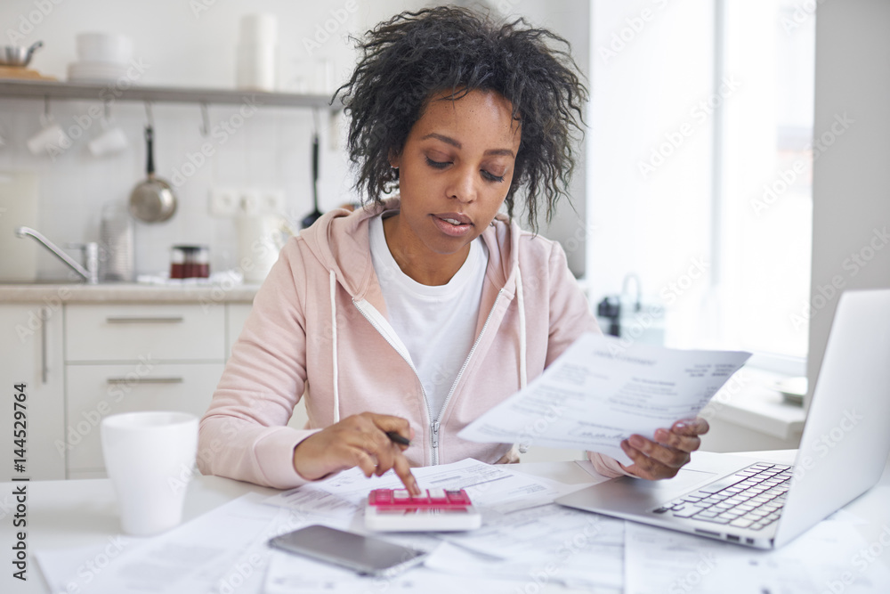 Financial literacy concept. Indoor shot of young single african american female student sitting at kitchen table, calculating expences, trying to analyze domestic dudget, thinking how to pay debt