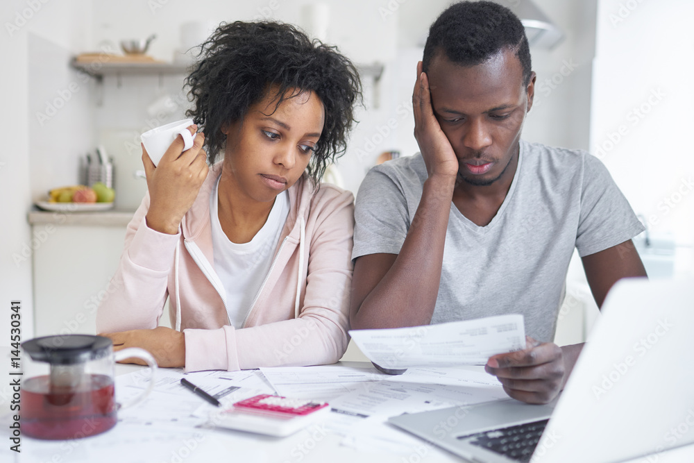 Depressed young African-American couple sitting at kitchen table with lots of papers. Family budget and debts concept. Husband holding notification from bank, trying to calculate domestic expenses