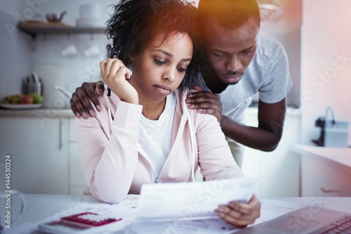 African man hugging wife, while she looking at laptop, using online banking app with, not able to pay off gas and electricity bills. Young couple sitting at kitchen table, facing financial problem photo