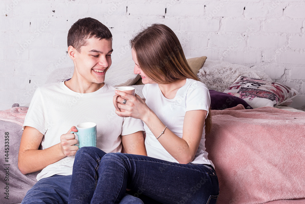 A loving couple sitting on a bed in the same T-shirts, smiling and looks at each other A loving couple sitting on a bed in the same T-shirts, smiling and looks at each other