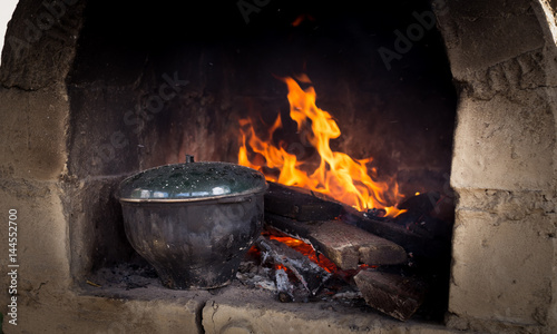 A fire burns in a stove. On coals a pot stands with a meal.