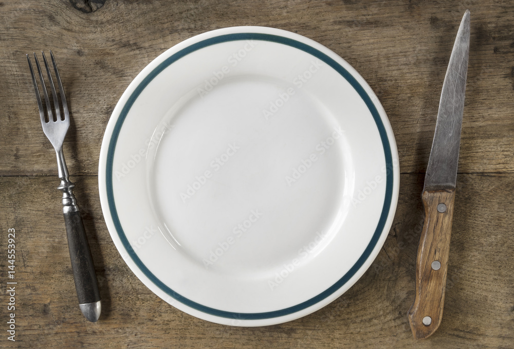 Small, empty plate, fork and knife on old wooden chopping board Small, empty plate, fork and knife on old wooden chopping board