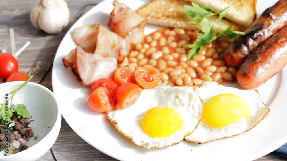 Full english breakfast - fried eggs, sausages, bacon, tomatoes, tomato beans, toasts - in white plate on wooden table. Slow pan. Full english breakfast - fried eggs, sausages, bacon, tomatoes, tomato beans, toasts - in white plate on wooden table. Slow pan.