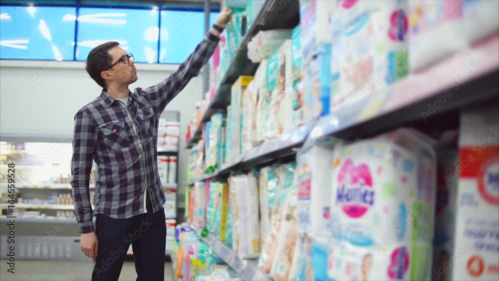 Man in the shopping center choosing goods. Choosing and inspecting the products for children diapers. Rows of shelves with goods for children. Concept of shopping in the mall. Man in the shopping center choosing goods. Choosing and inspecting the products for children diapers. Rows of shelves with goods for children. Concept of shopping in the mall.