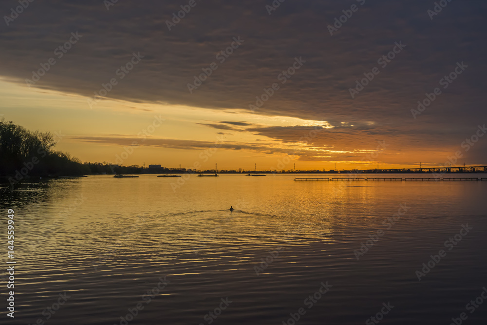 Deep orange sunrise under a stormy cloudy sky, reflected in calm lake water, bird swimming