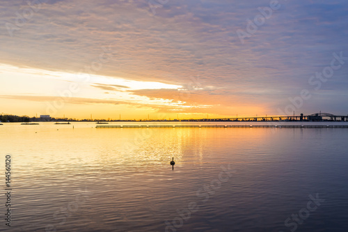 Fototapeta Naklejka Na Ścianę i Meble -  Deep golden sunrise over calm lake, silhouette of bird swimming in foreground, dramatic cloudscape