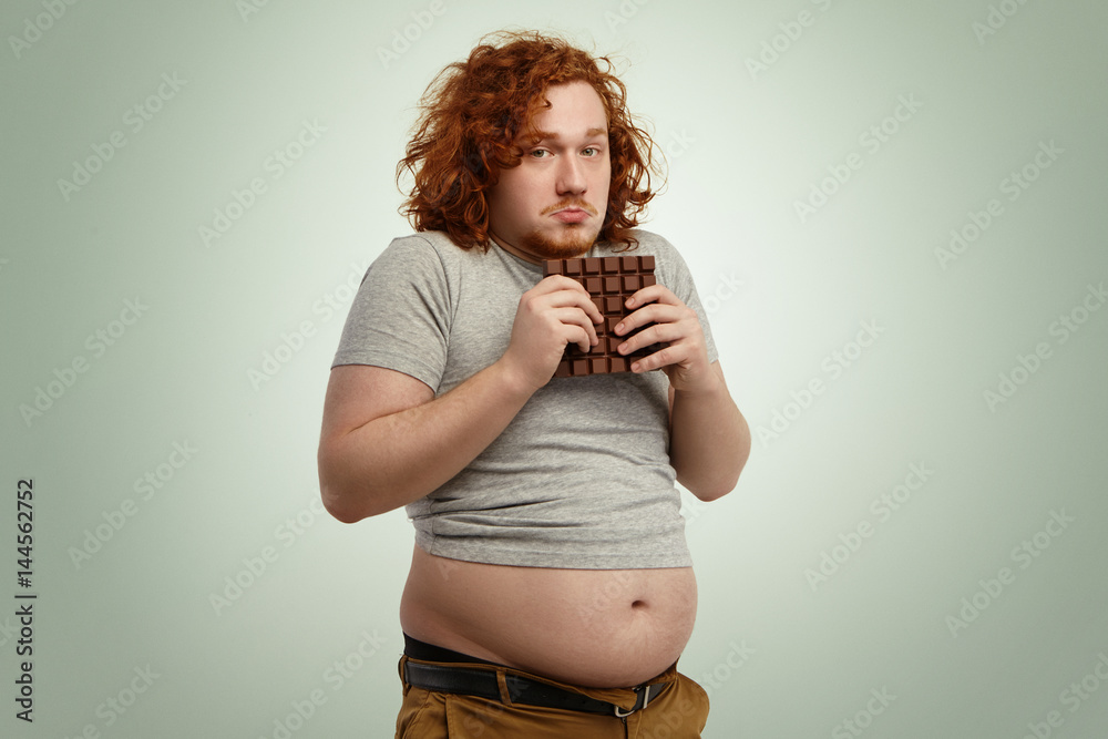 Overweight fat man with ginger curly hair looking indecisive and hesitant,  holding large bar of chocolate