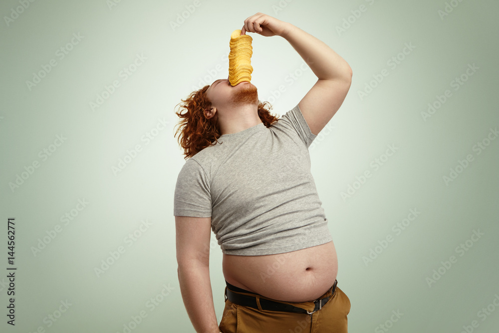Fat obese greedy redhead man throwing head back while trying to stuff big  pile of potato chips into his mouth, feeling hungry and impatient.  Overweight young male consuming junk food indoors Stock