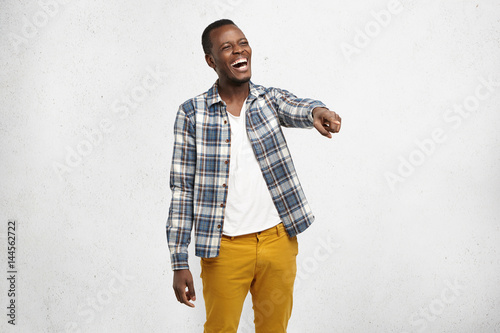 Body language, gestures. Portrait of charismatic dark-skinned student wearing checkered shirt, laughing at someone or something, pointing finger at subject, looking happy, cheerful and carefree