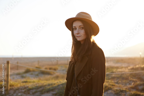 Outdoor portrait of beautifulCaucasian girl wearing fashionable coat and hat feeling carefree and peaceful, contemplating amazing morning view at seaside, looking at camera with charming smile
