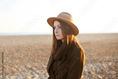 Attractive young European female dressed in stylish overclothes having nice walk along coastline on sunny day, came to the sea for contemplating sunset. Pretty girl in hat relaxing on sandy beach