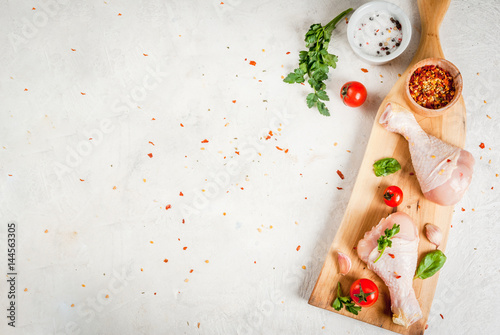 Raw chicken legs. Meat, a source of protein. On a cutting board, on a white stone table. With spices, herbs and tomatoes for cooking. Top view copy space