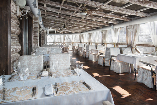 interior of the restaurant with panoramic windows and wooden walls. White tablecloths with gilding. Served tables with wine glasses