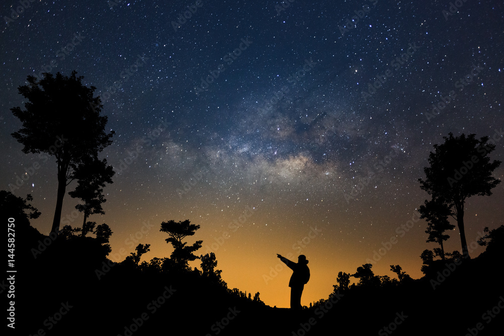 Obraz premium A Man is standing next to the milky way galaxy pointing on a bright star in forest, Long exposure photograph, with grain.