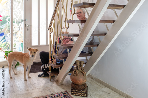 two senior men sit on staircase in hall, brown dog is standing beside photo