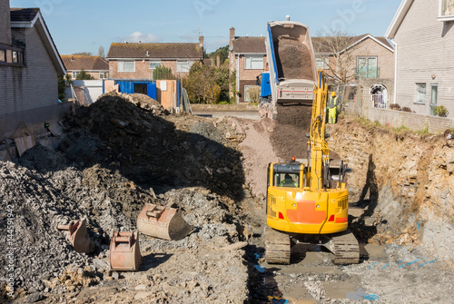 Excavator is digging a house foundation in a residential area while a dumper truck is unloading construction gravel, sand and crushed stones on the construction site.