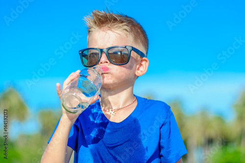 Portrait of a thirsty handsome boy in sunglasses drinking water on the desert tropical beach