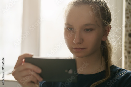 teen girl sitting near window with smartphone, toned photo with shallow focus