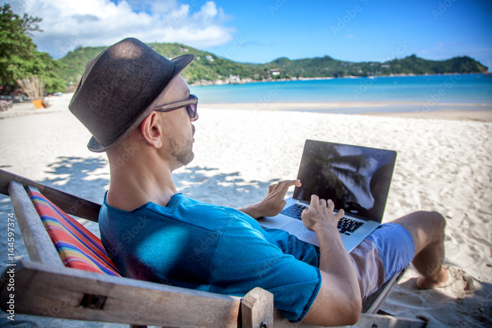 Young hipster man with laptop on tropical beach. Travel, vacation, internet, freelance job concept