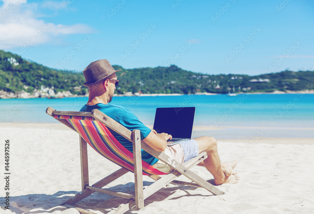 Young hipster man with laptop on tropical beach. Travel, vacation, internet, freelance job concept