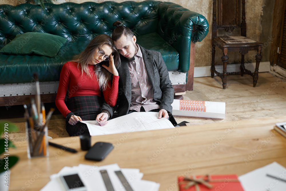 Tired colleagues with drawings sit on the floor near green sofa with working desk in front. Deadline concept. Stressed job for people. Responsible male and female team.