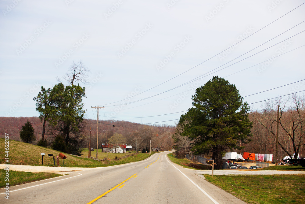 A curving blacktop highway through a village of buildings and old van tailers surrounded by forest of bare trees and evergreen trees in a countryside landscape
