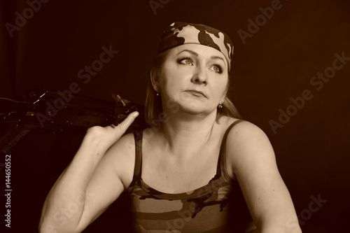 A middle aged, white, woman holds an AK74 automatic rifle against black photo