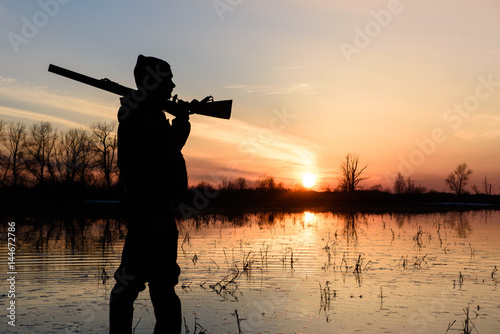 Silhouette of a hunter at sunset in the water with a gun. 