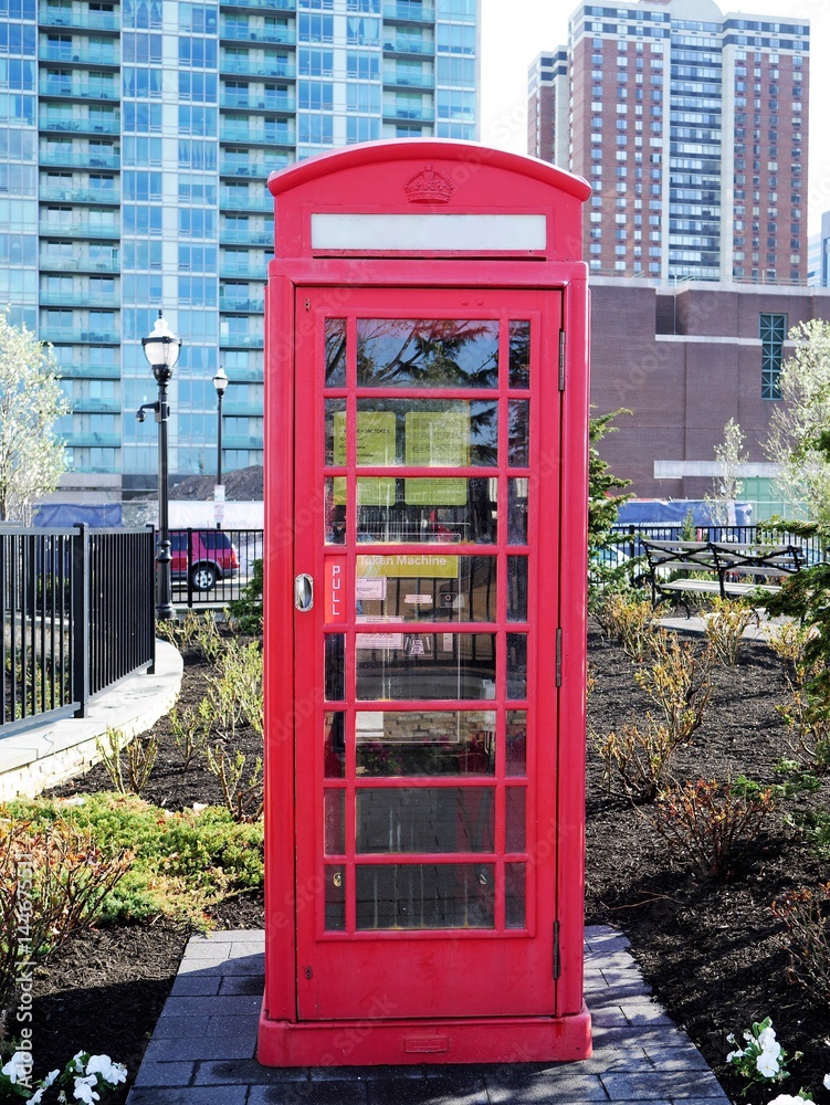Imitation classic British London red phone booth in a park with modern highrise building in the background Imitation classic British London red phone booth in a park with modern highrise building in the background