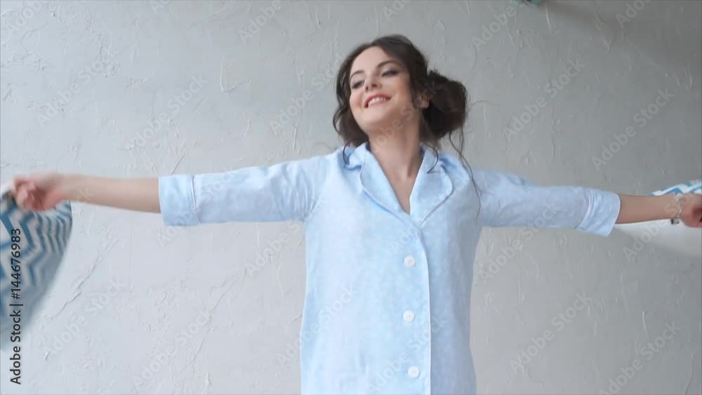 Young woman jumping on the bed in the bedroom, in her hands a few pillows. A happy beauty waving them in the air, she fools around and remembers her childhood Young woman jumping on the bed in the bedroom, in her hands a few pillows. A happy beauty waving them in the air, she fools around and remembers her childhood