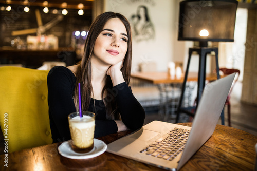 Young female with cute smile sitting with laptop in modern coffee shop interior during recreation time, charming happy woman student using laptop computer to prepare for the course work