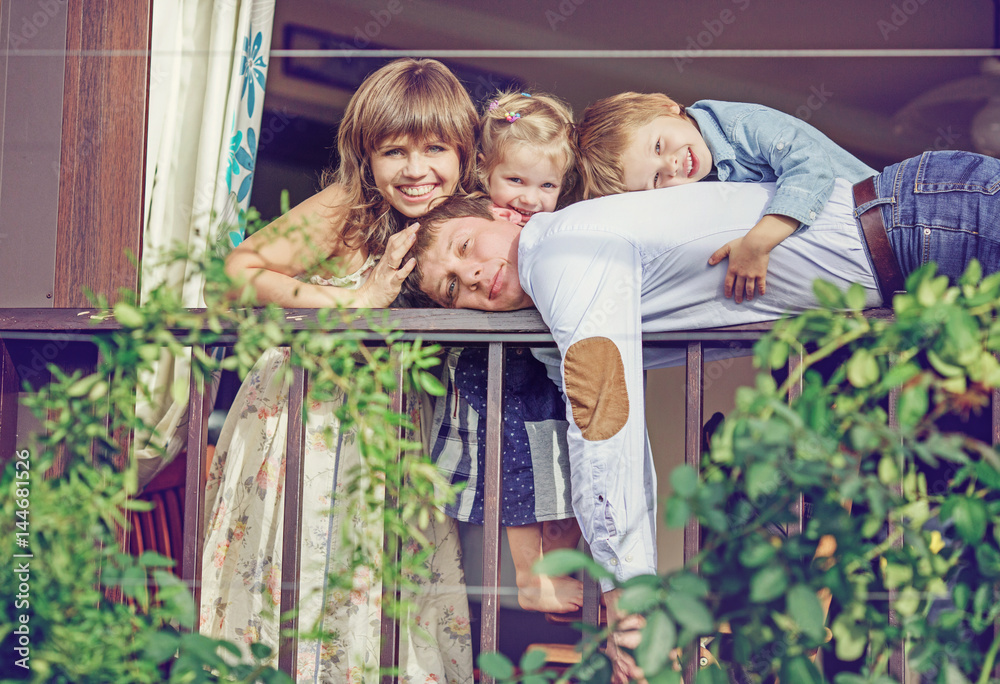 Big family peeks out from the balcony of their house. The man is lying on the railing, twined with green plants, and the children are hugging him Big family peeks out from the balcony of their house. The man is lying on the railing, twined with green plants, and the children are hugging him