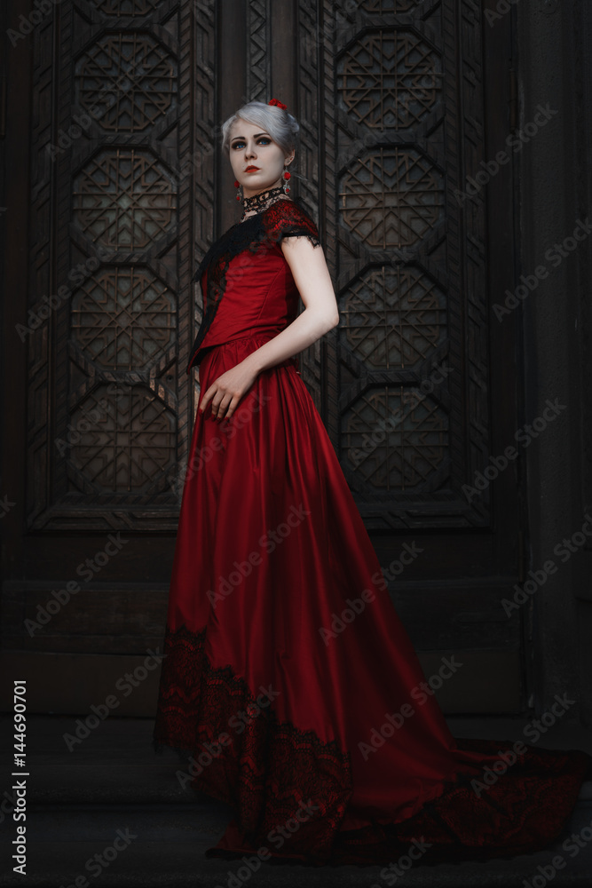 Woman in a red ball gown, she stands in front of a fairy-tale door to the castle.