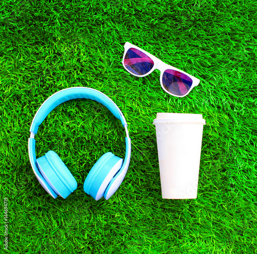 Blue headphones, coffee cup, sunglasses on a green grass textured background, top view, flat lay photo
