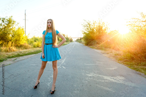 beautiful blonde girl in blue dress standing on a road at sunset background. photo