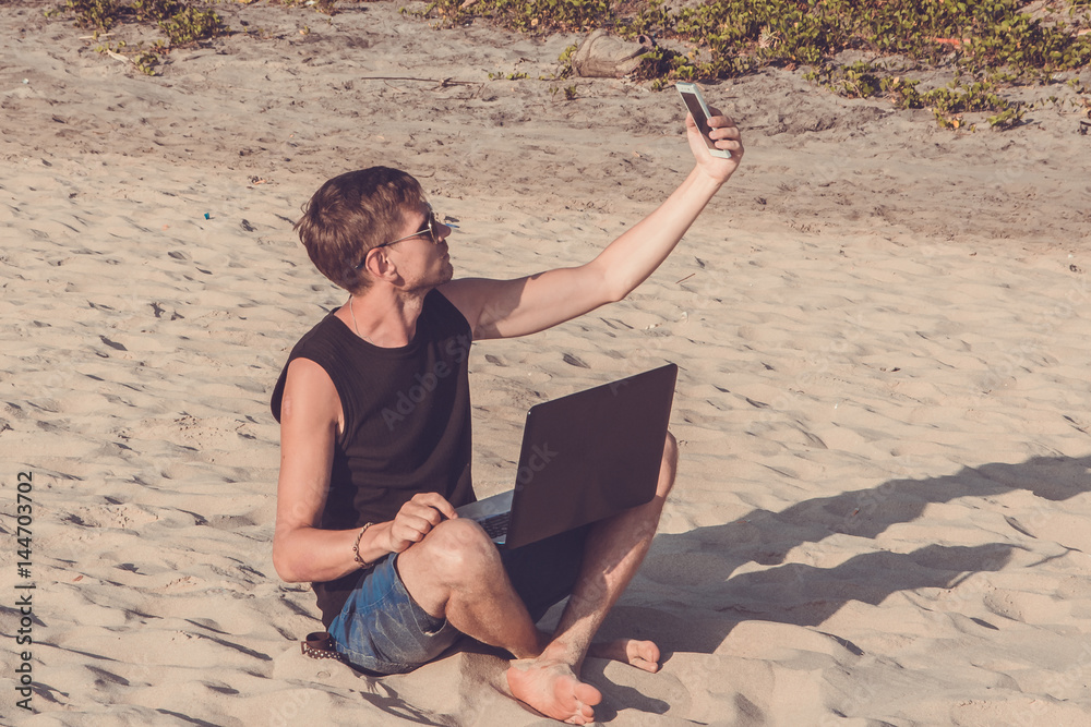 Young man with laptop computer and mobile phone on the beach. Downshifting and freelance concept.