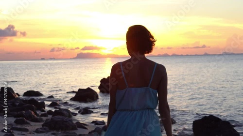 Happy young woman in a long dress dancing on the beach on a colourful sunset background
