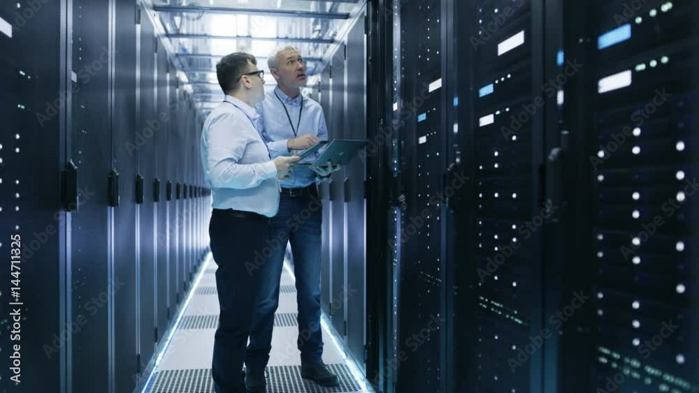 Two IT Engineers Standing in Working Data Center. They Use Laptop and Tablet Computer while Standing Beside Open Server Rack Cabinet.  Shot on RED EPIC-W 8K Helium Cinema Camera.