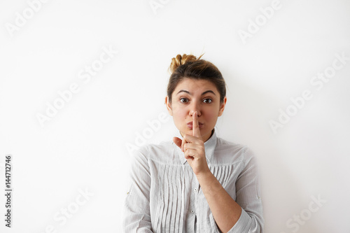 Keep silence! Portrait of pretty young businesswoman wearing in white striped shirt holding her finger near lips and making silence gesture asking to keep confidential information private