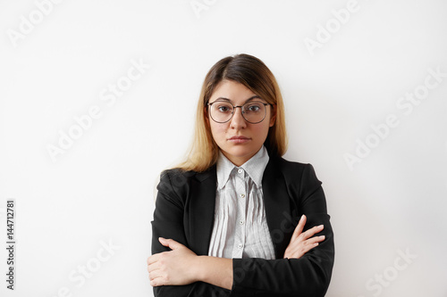 Portrait of successful young entrepreneur female in black suit and shirt wearing glasses looking confident and serious. Thoughtful young businesswoman standing staring at camera with folded arms