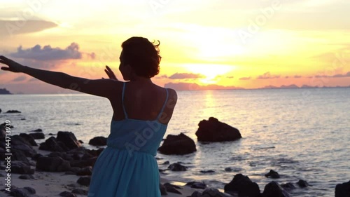 Happy young woman in a long dress dancing on the beach on a colourful sunset background
