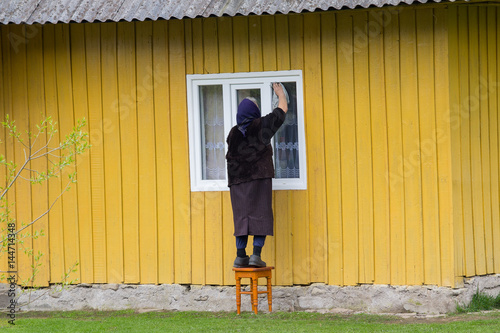 Elderly woman washes the window of a rural house. People