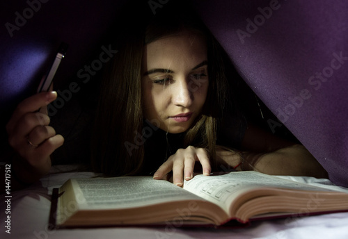 Young woman hiding under blanket and enrapt reading interesting book at nighttime. Girl lighting with the phone as a flashlight photo