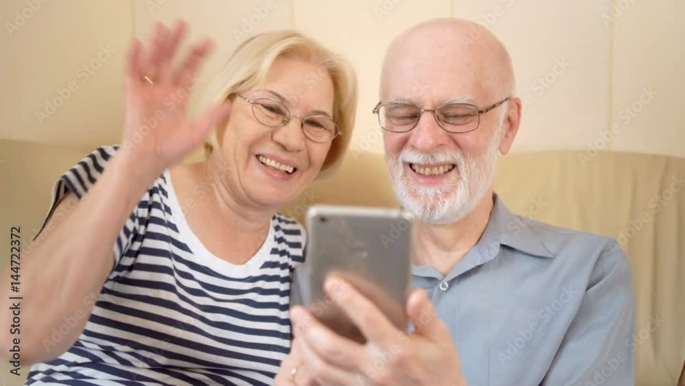 Cheerful good-looking senior couple sitting on sofa at home. Talking via Skype on smartphone Cheerful good-looking senior couple sitting on sofa at home. Talking via Skype on smartphone