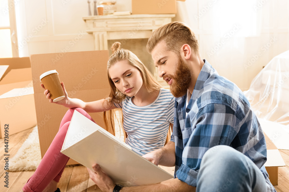 Young couple holding disposable coffee cup and album while sitting on floor in new house Young couple holding disposable coffee cup and album while sitting on floor in new house